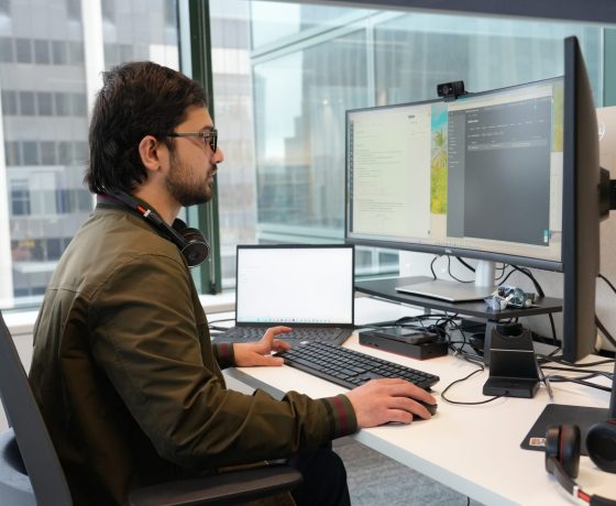 Zetta team member working at a dual-monitor desk with a laptop in the Perth office, city buildings through the window behind